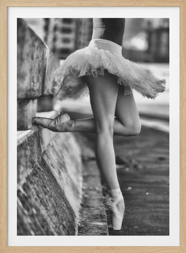 A black and white photograph focusing on the lower half of a ballerina in a tutu and pointe shoes. She stands en pointe with one foot on the ground, while the other foot rests against a rough, concrete curb, highlighting the contrast between delicate dance and an urban environment. Decor