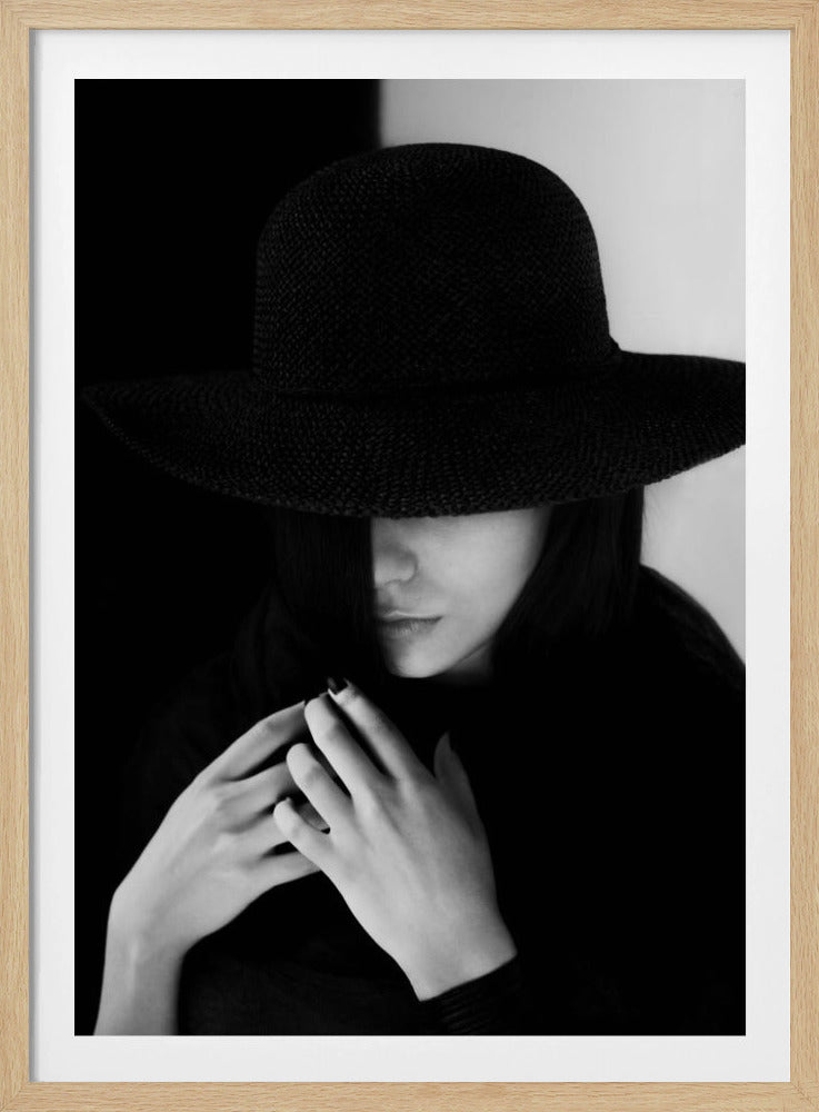 A mysterious black and white portrait of a woman wearing a large, black, wide-brimmed hat that casts a deep shadow over her eyes. Her hands are clasped at her chest, and the background is split vertically into black and white sections. Artwork