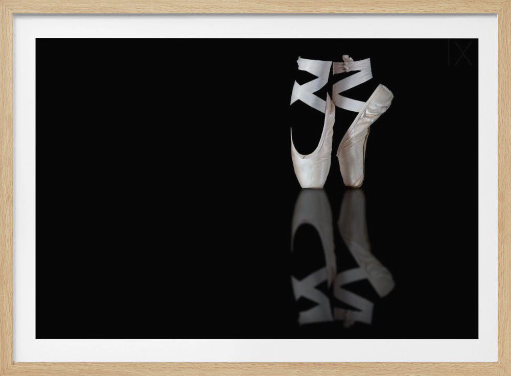 A pair of white satin pointe shoes with ribbons are positioned on their tips on the right side of the image against a solid black background, with a clear reflection beneath them on a glossy surface. The entire image is enclosed in a brushed silver frame. Print