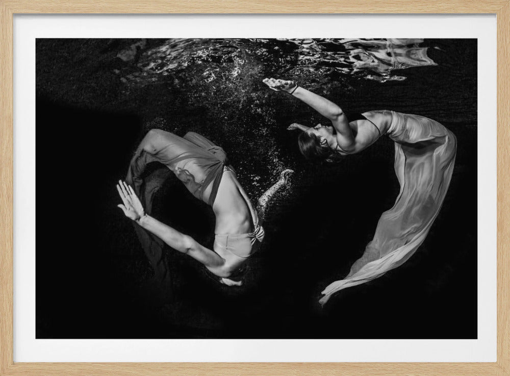 A striking black and white underwater photograph of two women in flowing fabric, their bodies arched gracefully in the dark water. One woman arches backwards towards the rippling surface, while the other curls forward, creating a dynamic, almost yin-yang composition against the deep black background. The photo is enclosed in a silver frame. Decor