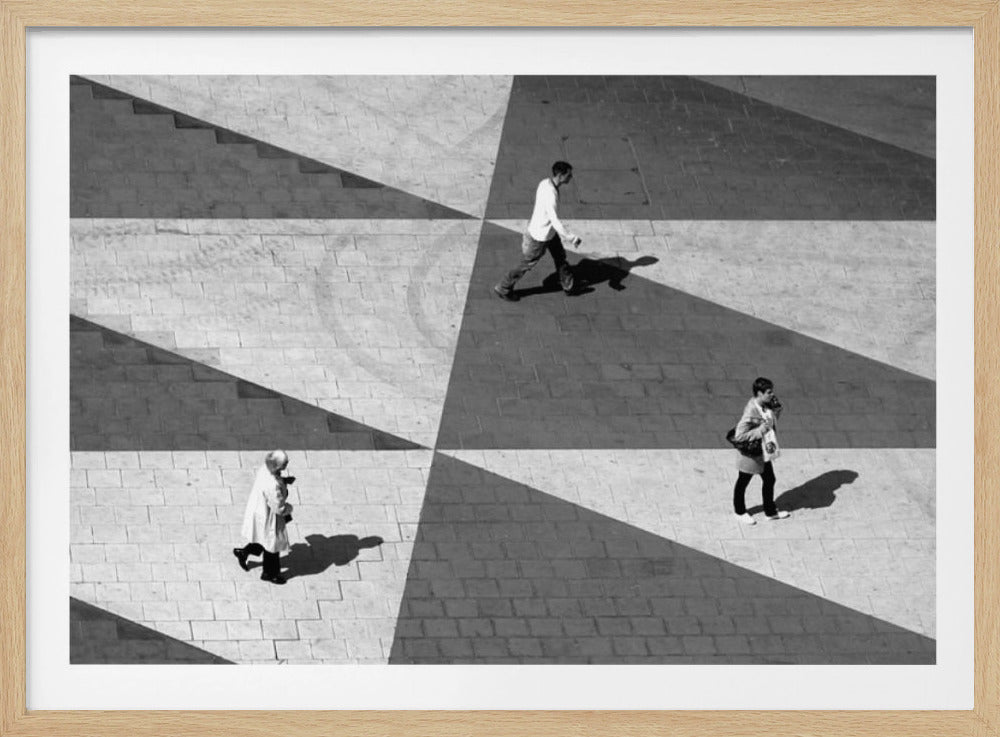 A high-angle, black and white photograph capturing three people walking across a paved plaza, their paths intersecting with a dramatic geometric pattern of light and shadow. The strong sunlight creates large, sharp-edged triangles of shade, isolating each figure in the composition, which is enclosed in a silver frame. Decor