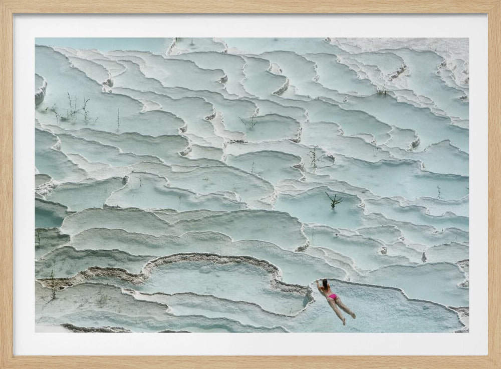 An aerial view of a woman in a pink bikini swimming in the stunning, terraced, milky-blue thermal pools of Pamukkale, Turkey. The white travertine formations create a unique, layered landscape, and the entire image is enclosed in a silver frame. Artwork