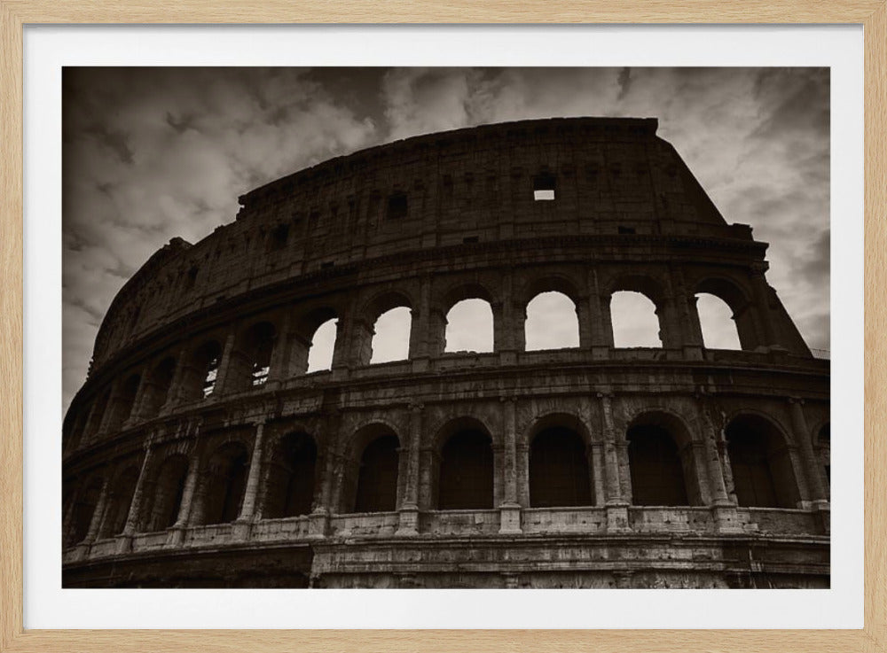 A low-angle, sepia-toned photograph of the Roman Colosseum against a dramatic, cloudy sky. The image focuses on the curved, weathered stone walls and the multiple tiers of arched windows of the ancient amphitheater. The photograph is presented in a silver frame. Decor