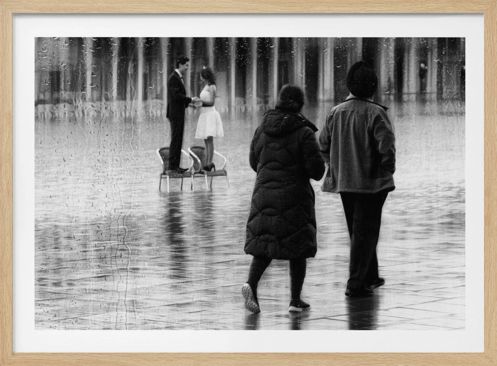 A black and white photograph, seen through a rainy window, captures a surreal city scene. In the background, a couple in formal wear stands on chairs in a flooded plaza, holding hands. In the foreground, the backs of two people in winter coats are seen as they walk on the wet pavement, observing the couple. Decor
