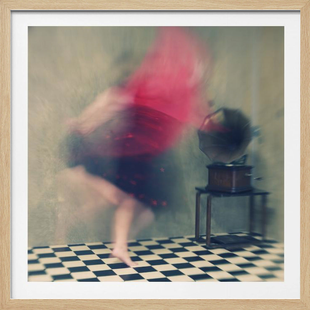 A motion-blurred, artistic photograph in a light wood frame showing a woman dancing barefoot on a black and white checkered floor. She wears a bright red top and a dark skirt, both blurred by her movement. Next to her stands a vintage gramophone on a small table, against a muted, textured wall, creating a dreamy, nostalgic atmosphere. Print