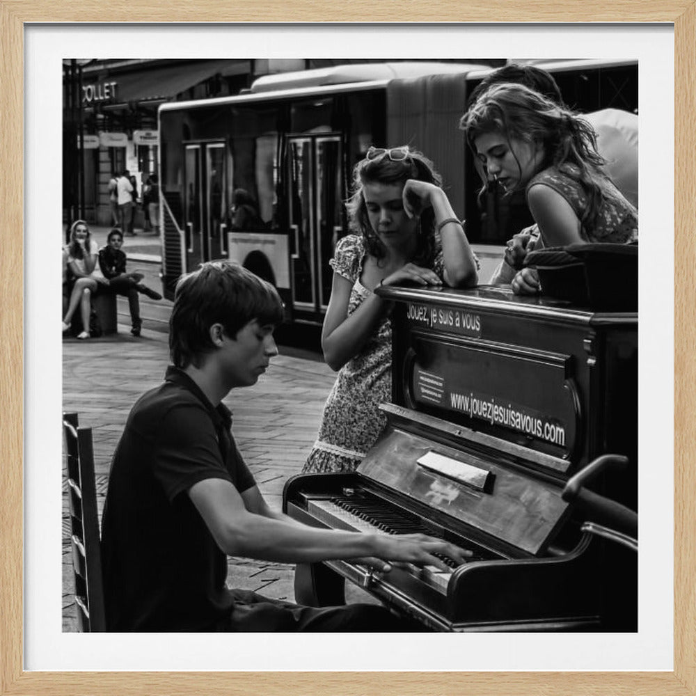 A black and white photograph in a light wood frame capturing a candid street scene. A young man plays an upright piano on a sidewalk while two young women lean on the instrument, watching him intently. The background shows a city street with a bus and other people. Wall Art
