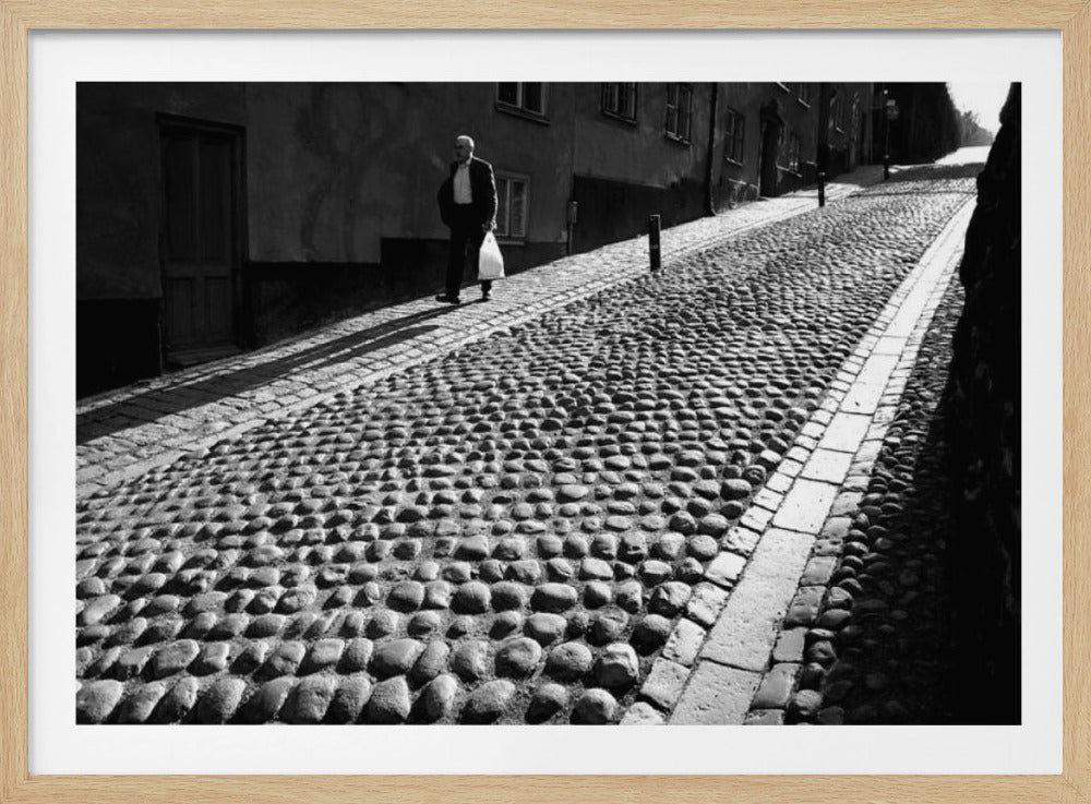 A framed, black and white photograph capturing an elderly man in a suit walking up a steep cobblestone street. The man carries a white bag and casts a long shadow behind him in the harsh sunlight. The street is lined with old, multi-story buildings, creating a classic European city scene. Poster