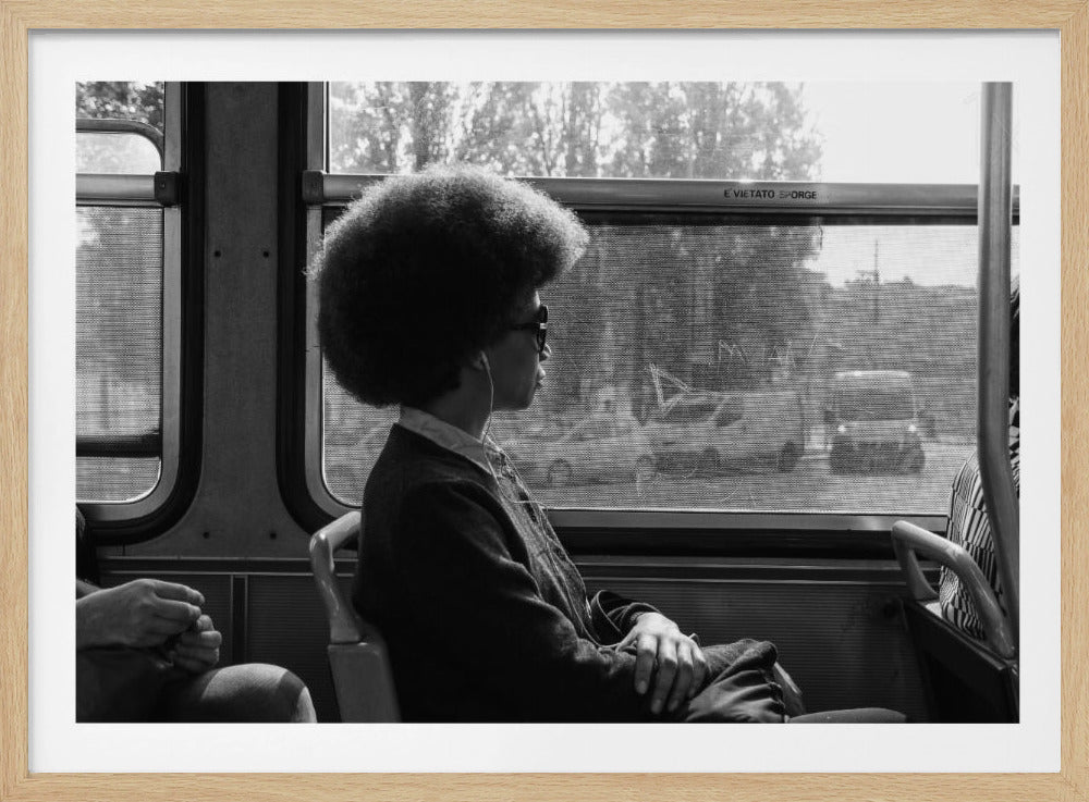 A black and white framed photograph of a person with a large afro hairstyle, wearing glasses and earphones, sitting in profile on a bus and looking thoughtfully out the window. Decor