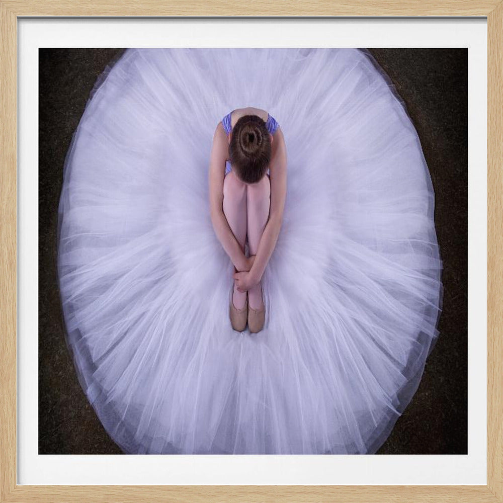 An overhead photograph of a young ballerina curled up on a dark floor, her massive white tutu fanned out in a perfect circle around her. The image is presented within a light wood frame. Print