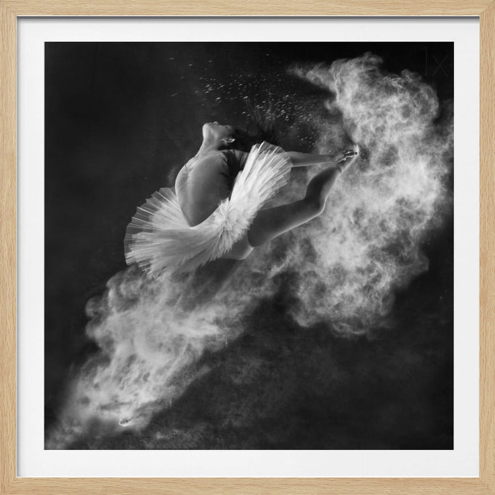 A dramatic black and white overhead photograph of a ballerina in a white tutu, captured mid-air in an arched pose. She is surrounded by an explosive cloud of white powder against a dark background. The entire artwork is presented in a light-colored wooden frame. Artwork