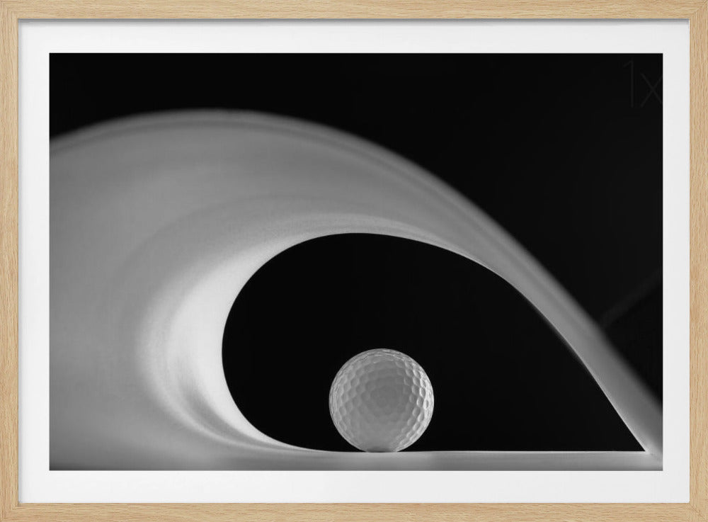 A dramatic black and white still life photo featuring a golf ball set against a stark black background. A large, abstract white shape swirls gracefully over the ball, creating a dynamic, wave-like composition. The image is presented within a silver frame. Decor