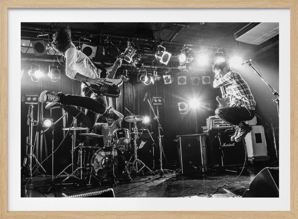 A high-energy, black and white photograph of a rock band performing live. The bassist and guitarist are frozen in mid-air as they jump, while the drummer plays in the background, all under bright stage lights. Print