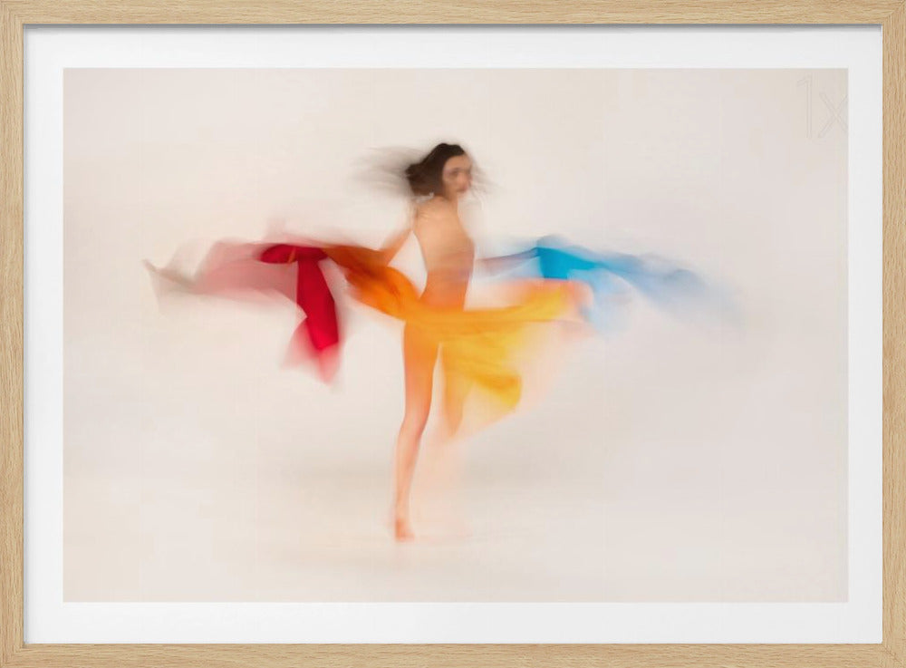 A long-exposure photograph of a woman dancing, creating an abstract image with motion blur. Her figure is softly defined against a plain white background, and she is enveloped by vibrant, flowing trails of color in red, orange, and blue, suggesting graceful movement. The artwork is displayed in a thin silver frame. Artwork