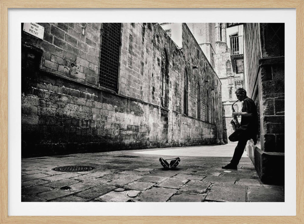 A black and white photograph of a man playing the saxophone while leaning against a stone wall in a narrow, cobblestone European alley. An open case for tips rests on the ground in a patch of sunlight. Poster