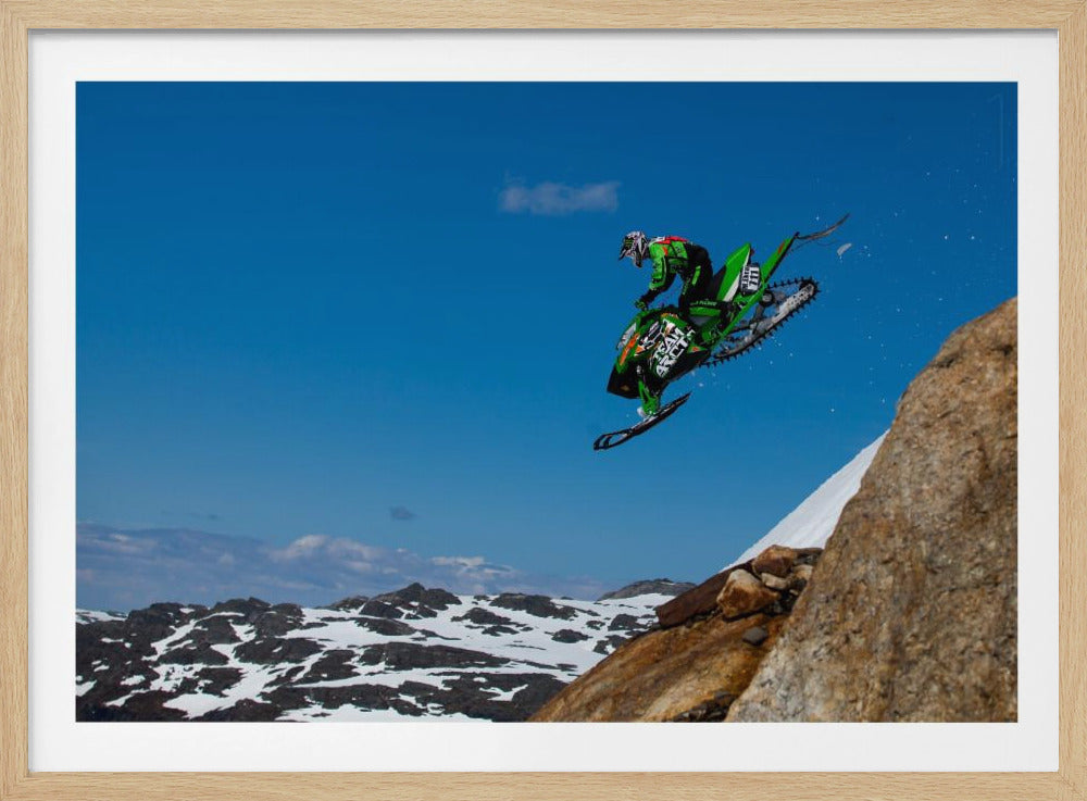 A dynamic action shot of a person in full gear launching a green snowmobile into the air off a rocky, snow-covered cliff, set against a backdrop of a vivid blue sky and distant snowy mountains. The image is enclosed in a silver frame. Wall Art