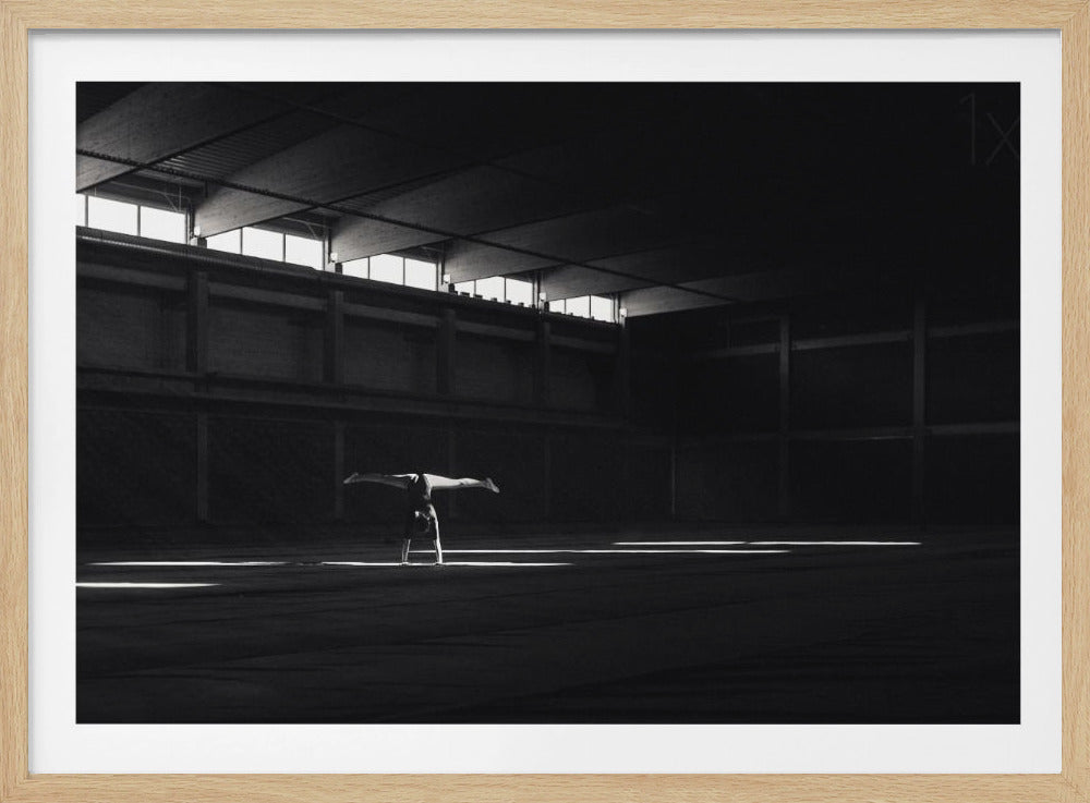 A dramatic black and white photograph of a gymnast holding a handstand with legs split, perfectly silhouetted in a single beam of light within a vast, dark industrial hall. Print