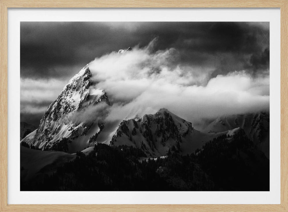 A dramatic black and white photograph of a snow-covered mountain peak being enveloped by thick, swirling clouds under a dark, stormy sky. The rugged slopes and sharp ridges of the mountain are highlighted by the snow, while a dark forest sits at the base. The entire image is presented within a textured silver frame. Print