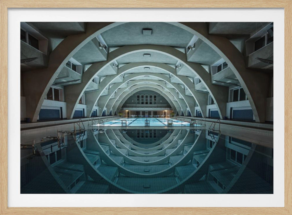An atmospheric photograph of an empty indoor swimming pool with remarkable Brutalist-style architecture. A series of large, repeating concrete arches span the length of the pool, creating a tunnel-like effect. The arches and lights are perfectly reflected in the still, blue water, forming a symmetrical, eye-like shape. Decor