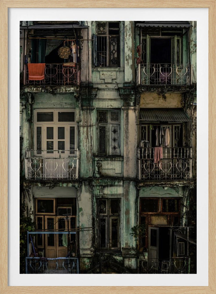 A tightly framed, symmetrical photograph of a decaying multi-story apartment building facade. The pale green paint is peeling and stained with dark grime, revealing the building's age. Each of the nine visible sections features a balcony with ornate ironwork or a window, showing glimpses of daily life through laundry hanging out to dry and dimly lit interiors. Poster