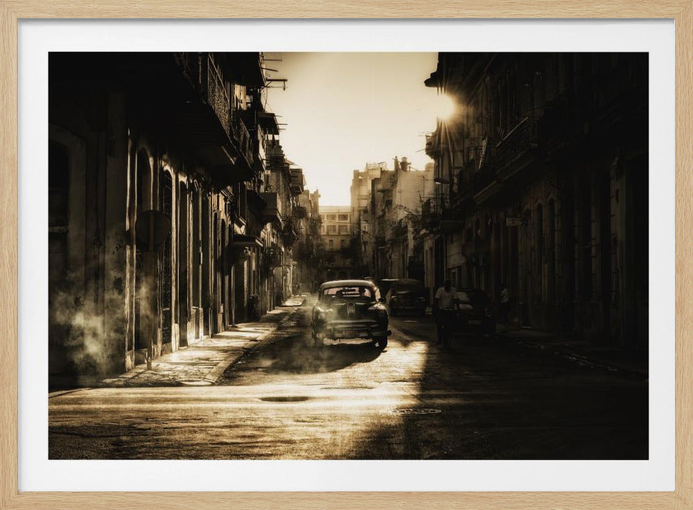 A sepia-toned, atmospheric photograph of a classic car driving down a narrow street in an old city. The sun hangs low, casting long shadows and illuminating smoke or dust in the golden air. Artwork