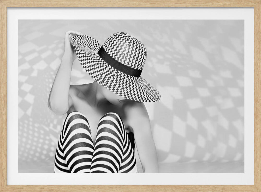 A black and white artistic photograph of a woman sitting down, her face obscured by a large, wide-brimmed sun hat with a black and white chevron pattern. She is wearing black and white striped leggings and holding the brim of her hat with one hand. The image is set against a plain background with patterned shadows and is presented within a silver frame. Artwork