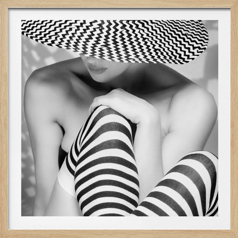 A framed, black and white photograph of a woman wearing a large hat with a chevron pattern that hides her eyes. She is also wearing black and white striped stockings and is posed hugging her knees. The photo has a strong graphic quality due to the contrasting patterns. Decor