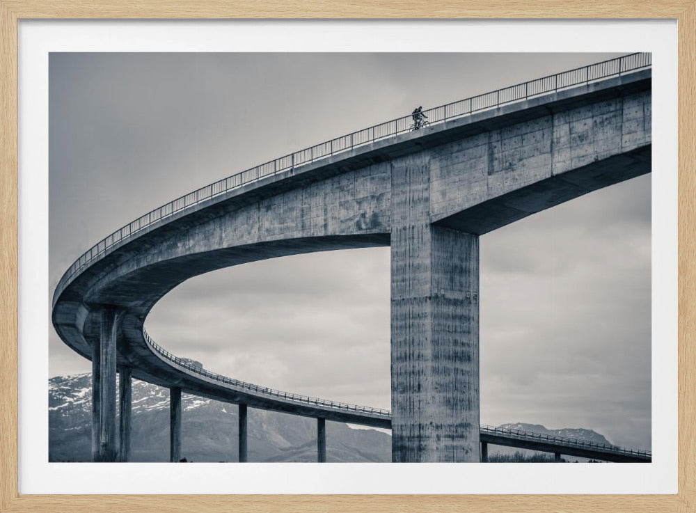 A black and white photograph of a lone cyclist riding across the top level of a massive, curving concrete bridge, which sweeps across the frame. In the background, a lower level of the bridge and distant mountains are visible under a cloudy sky. Decor