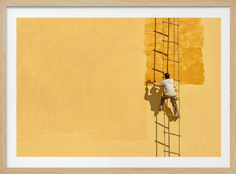 A man viewed from behind stands on a rope ladder, painting a large, bright yellow wall a slightly darker shade of yellow, casting a stark shadow in the bright sun. The image is presented within a silver frame. Decor