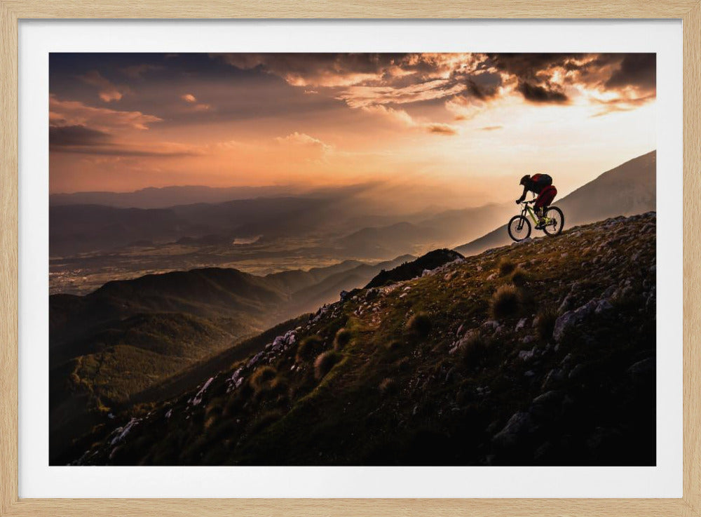 A lone mountain biker is silhouetted against a breathtaking sunset as they navigate a steep, rocky ridge. Below them, layers of mountains and a vast valley stretch into the distance under a dramatic, cloudy orange sky. Artwork