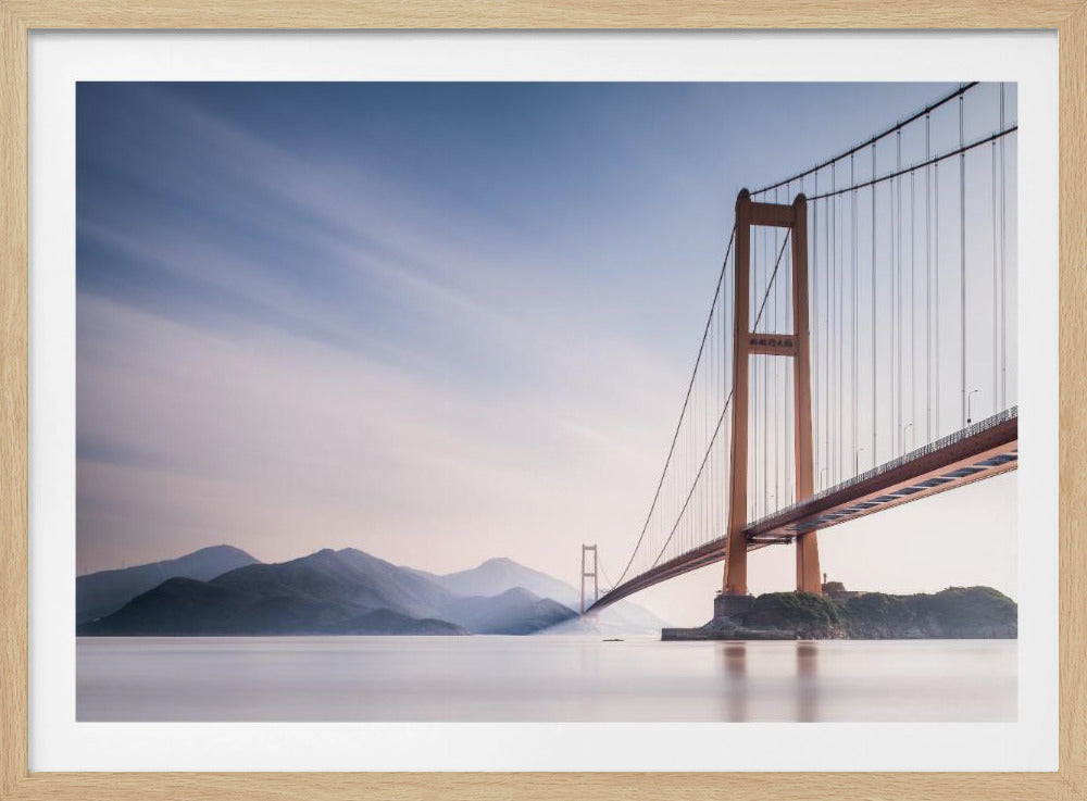 A serene long-exposure photograph of a golden suspension bridge stretching over calm, milky water, with hazy mountains visible in the distance under a soft blue and pink sky. Print