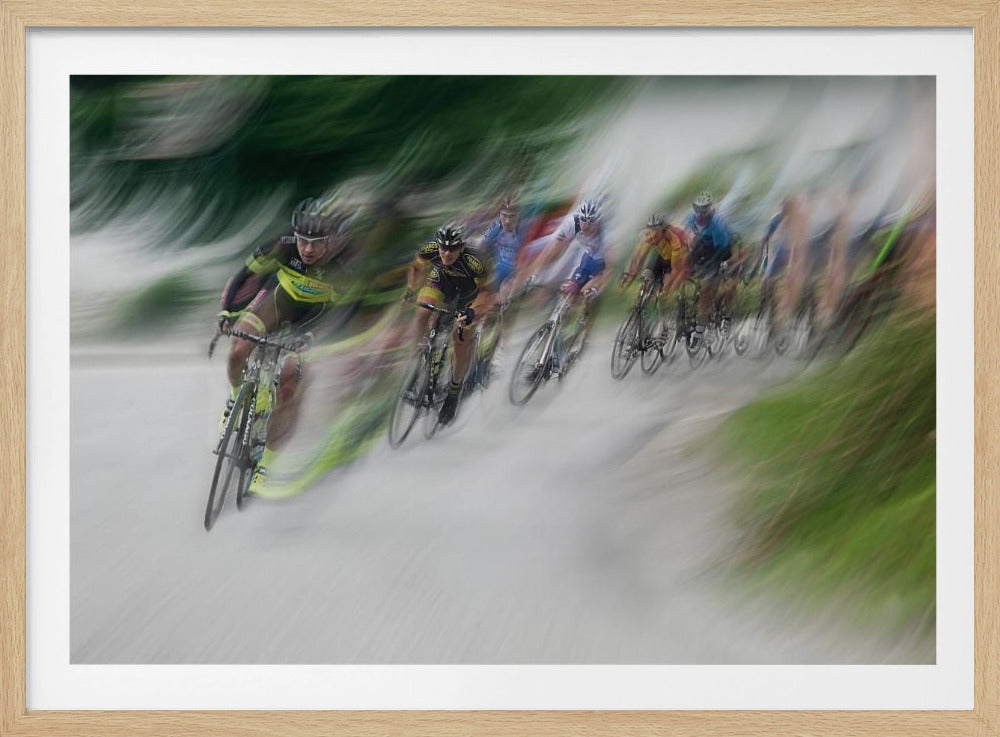 A dynamic, motion-blurred photograph of a group of cyclists in a race, captured with a slow shutter speed to create streaks of color and a sense of high speed. The lead cyclist in a yellow and black jersey is the most defined figure, leaning into a turn on a grey road flanked by blurry green foliage. The entire image is enclosed in a silver frame. Decor