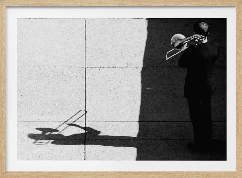 A high-contrast, black and white photograph of a man in a suit playing a trombone, seen from behind. He stands in a stark shadow that cuts across a sunlit concrete sidewalk, where a long shadow of him and his instrument is cast. Artwork
