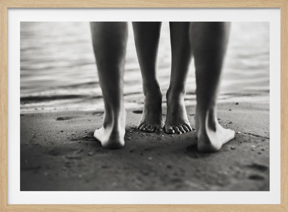 A framed, black and white photograph showing a low-angle view of two people's bare feet and lower legs standing on a wet sandy beach. The central pair of feet is in focus, while another person's legs in the foreground are blurred, creating depth. The water's edge is visible in the background. Artwork