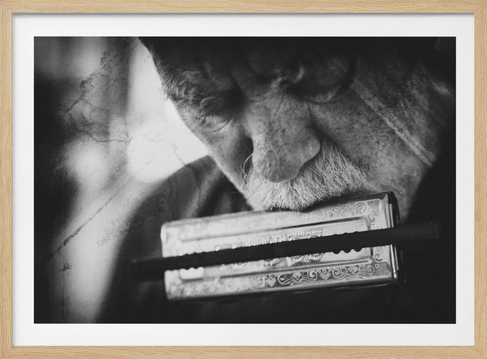 A close-up, black and white, vintage-style photograph of an elderly man with a white mustache playing a harmonica. His eyes are downcast in concentration, and the image is tightly framed, focusing on his face and the instrument, all within a silver border. Decor