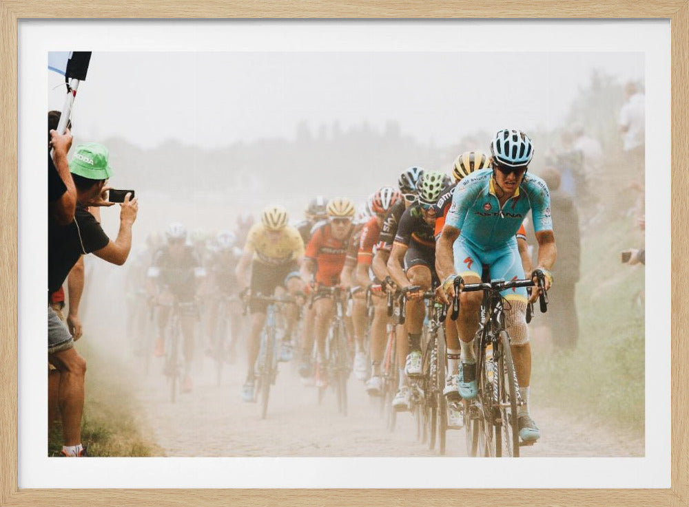 A framed photograph of a peloton of professional cyclists racing down a dusty dirt road. The lead cyclist is in sharp focus, wearing a bright turquoise uniform and a helmet, with other racers in yellow and red jerseys blurred behind him. A spectator on the left is taking a photo with a smartphone. Poster