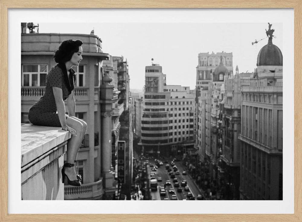 A framed, black and white photograph of a woman in a vintage-style polka dot dress and high heels sitting on the ledge of a tall building. She gazes down thoughtfully at the bustling city street and architecture below. Decor