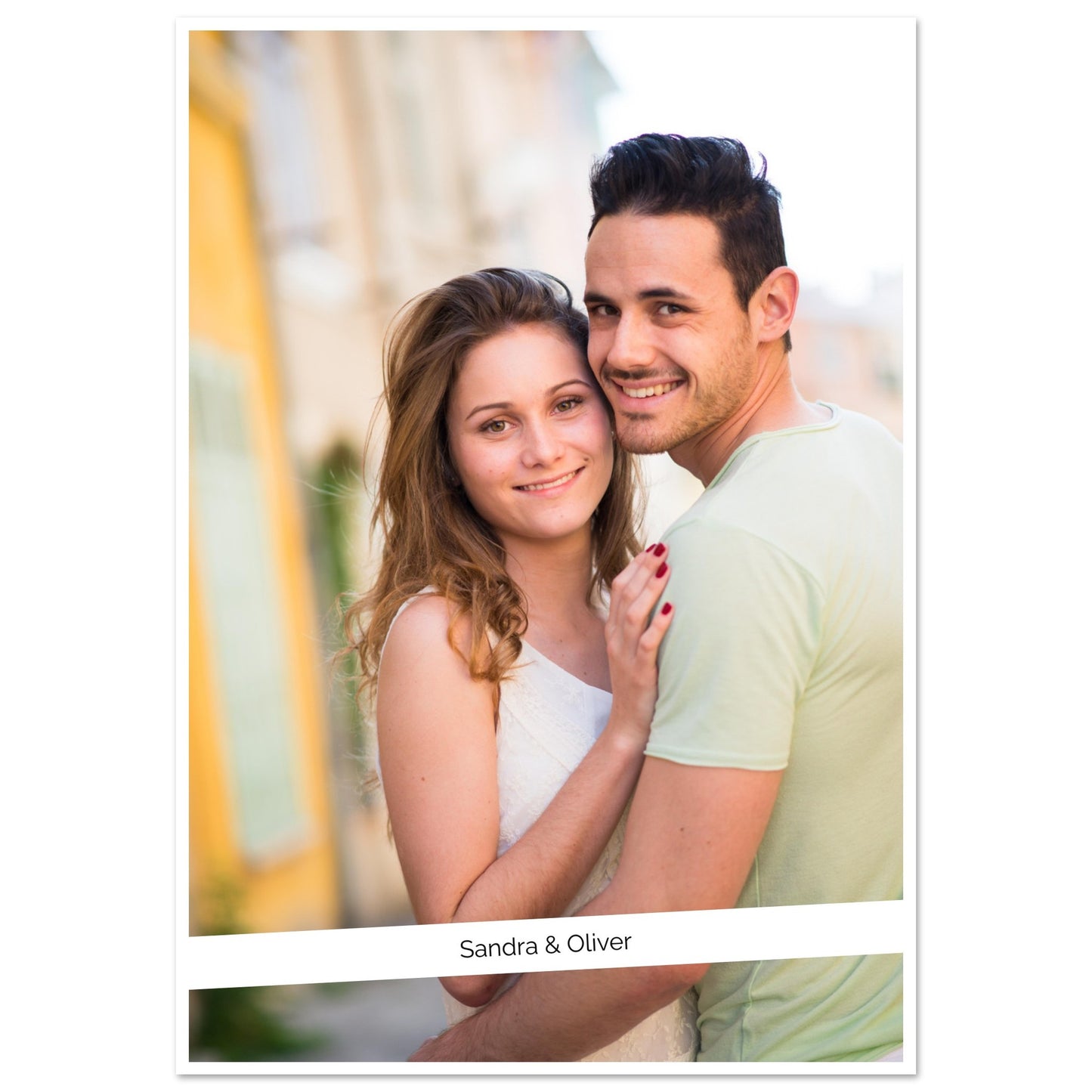 A portrait-style photograph of a smiling young couple, Sandra and Oliver, posed outdoors. The woman, with long brown hair and a white top, embraces the man, who is wearing a light green shirt. The photo is set against a brightly lit, soft-focus background of a street with yellow buildings. The image is framed with a white border and their names are printed in a simple font on a white bar at the bottom. Wall Art