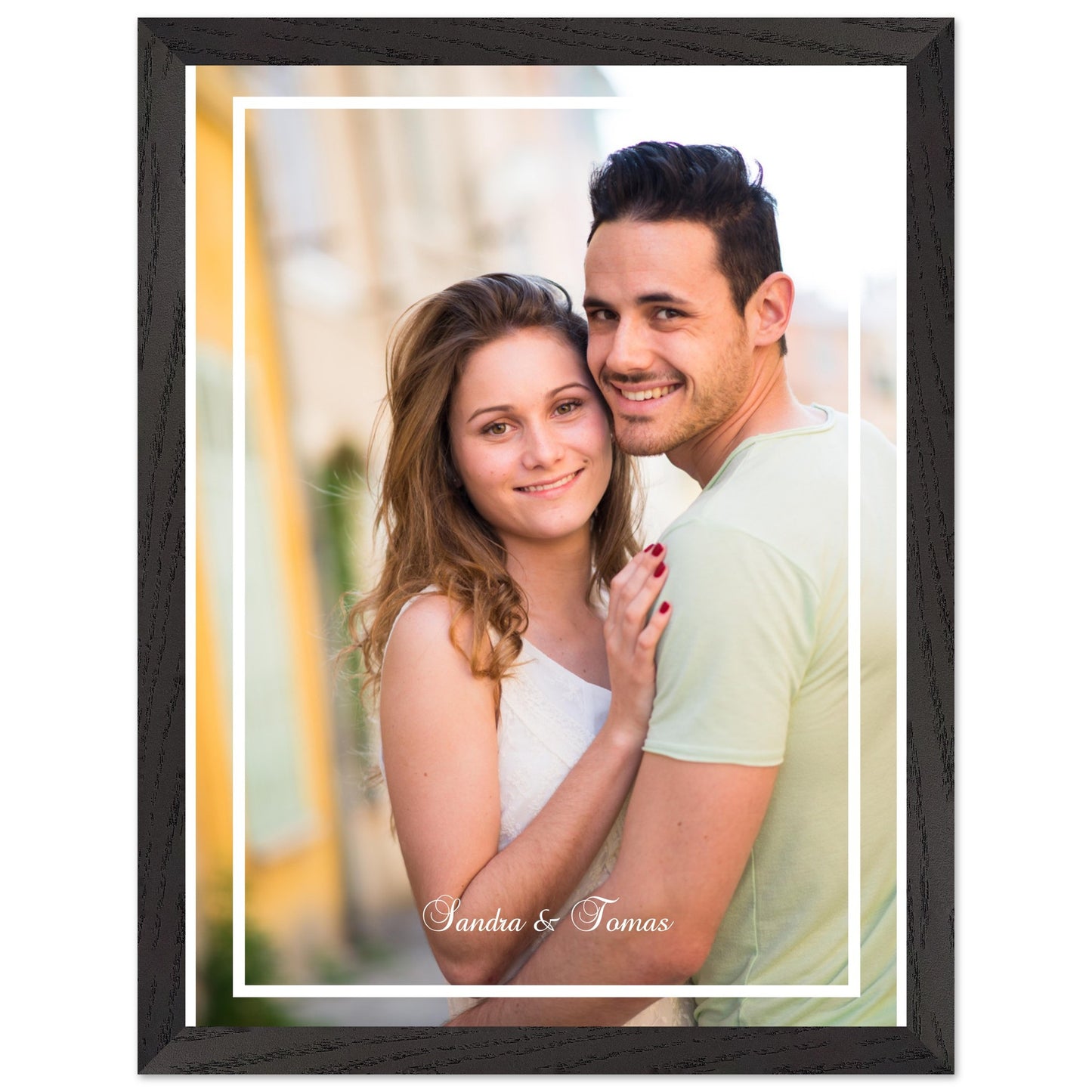A personalized framed photo of a smiling young couple, Sandra and Tomas, embracing. The vertical portrait has a white border and is set within a black wood-grain frame. The woman has long brown hair and a white top, while the man wears a light green shirt. Print