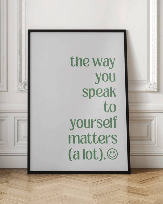 A minimalist poster with a black frame featuring the quote 'the way you speak to yourself matters (a lot).' in a sage green, lowercase serif font on a white background. The text is aligned to the right, with a small smiley face icon at the end. Decor