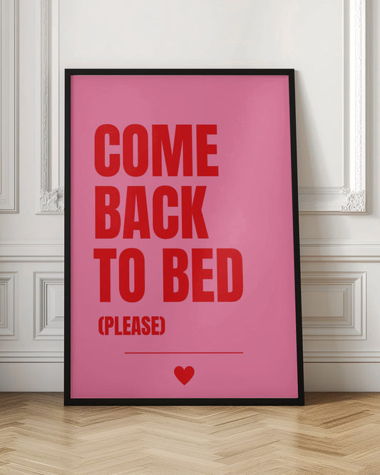 A framed poster with a pink background and bold red text. The text reads 'COME BACK TO BED' in all caps, with the words stacked. Below, in smaller letters, it says '(PLEASE)'. A small red heart is centered at the very bottom above a horizontal line. Artwork