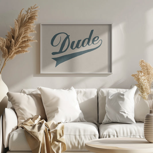 A framed artwork featuring the word 'Dude' in a retro, dark teal cursive script on a cream-colored canvas. The script has a long, swooping tail underline, reminiscent of a vintage sports logo, and is enclosed in a simple silver-gray frame. Artwork