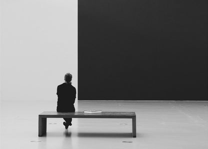 A black and white photograph of a person seen from behind, sitting alone on a wooden bench in a minimalist art gallery. They face a large, starkly divided wall, half white and half black. The scene is presented within a silver picture frame. Poster