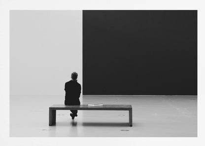 A black and white photograph of a person seen from behind, sitting alone on a wooden bench in a minimalist art gallery. They face a large, starkly divided wall, half white and half black. The scene is presented within a silver picture frame. Poster