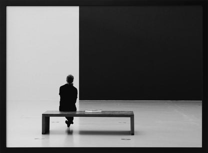 A black and white photograph of a person seen from behind, sitting alone on a wooden bench in a minimalist art gallery. They face a large, starkly divided wall, half white and half black. The scene is presented within a silver picture frame. Poster