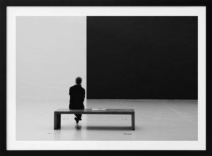 A black and white photograph of a person seen from behind, sitting alone on a wooden bench in a minimalist art gallery. They face a large, starkly divided wall, half white and half black. The scene is presented within a silver picture frame. Poster