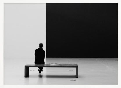 A black and white photograph of a person seen from behind, sitting alone on a wooden bench in a minimalist art gallery. They face a large, starkly divided wall, half white and half black. The scene is presented within a silver picture frame. Poster