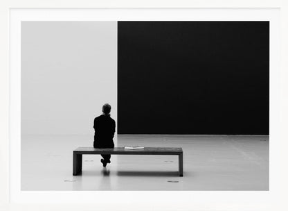 A black and white photograph of a person seen from behind, sitting alone on a wooden bench in a minimalist art gallery. They face a large, starkly divided wall, half white and half black. The scene is presented within a silver picture frame. Poster