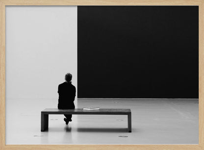 A black and white photograph of a person seen from behind, sitting alone on a wooden bench in a minimalist art gallery. They face a large, starkly divided wall, half white and half black. The scene is presented within a silver picture frame. Poster
