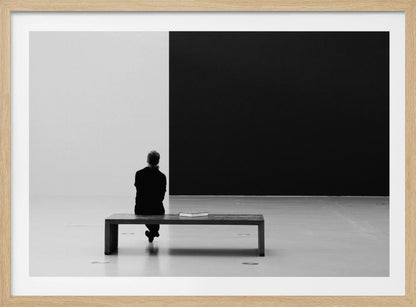 A black and white photograph of a person seen from behind, sitting alone on a wooden bench in a minimalist art gallery. They face a large, starkly divided wall, half white and half black. The scene is presented within a silver picture frame. Poster