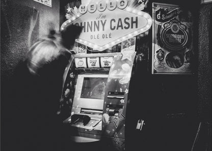 A grainy, black and white photograph of a person blurred by motion standing in front of a Johnny Cash themed slot machine. A lit marquee sign above the machine reads "HELLO I'm JOHNNY CASH OLÉ OLÉ". The setting appears to be a dark bar or casino. Wall Art